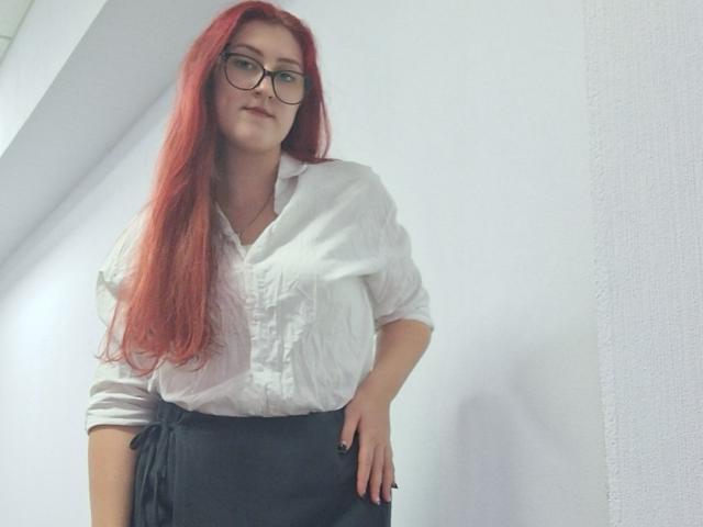 Triziya with long red hair, glasses, white button-up shirt, black skirt, hand on hip, looking down, slightly smiling, white wall in background.