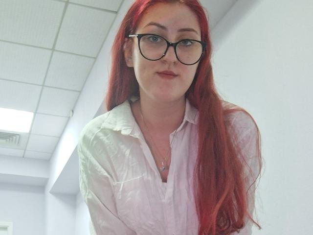 Triziya has long red hair, glasses, and a white button-up shirt, showing a slight smile with a small nose ring and a necklace in a bright room.