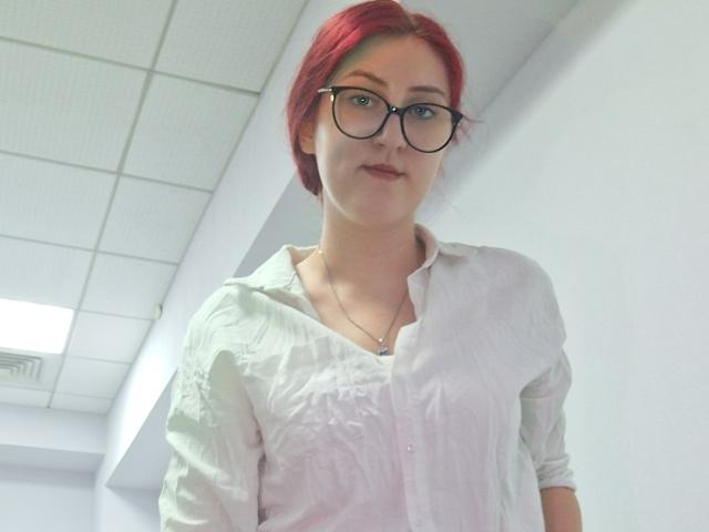 Triziya with red hair, glasses, and a white button-up shirt, standing in a bright room, showing her tits slightly through the shirt. She's looking down at the camera.