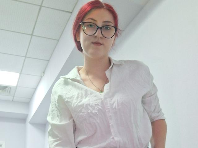 Triziya with red hair, glasses, and white button-up shirt, her tits slightly visible under the fabric, standing in a bright room, looking down at the camera.