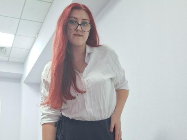 Triziya has long red hair, glasses, and wears a white button-up shirt with black pants, standing in a white-walled room, looking slightly down with a relaxed expression.