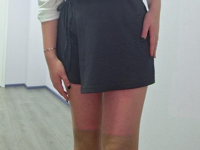 Triziya's black skirt, white top, black nails, bracelet, and sunburned thighs, standing with hands in skirt pockets. Her skirt is short, showing her sunburned ass.