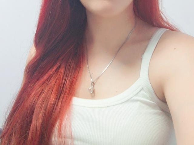 Triziya with red hair, white tank top, and silver necklace, showing her tits and a small pendant, upper chest and face partially visible.