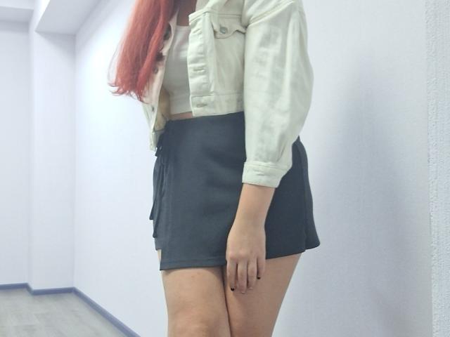 Triziya with long red hair, wearing a white jacket and black skirt, standing in a hallway, showing her ass and a hint of her pussy.
