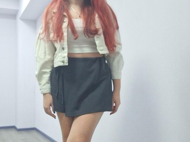 Triziya with long red hair, white crop top, pale green jacket, and black skirt, standing with legs slightly apart, showing a small bit of her ass.