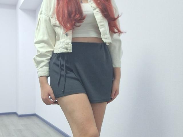 Triziya with red hair, white crop top, and black shorts, wearing a beige jacket, standing in a white-walled room, showing her ass and a slight tan on her thighs.