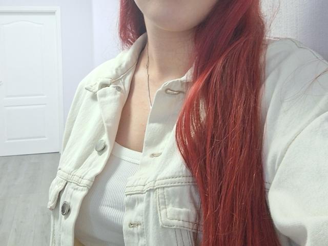 Triziya with red hair, wearing a light green denim jacket over a white ribbed top, visible chest, subtle necklace, door in the background, wooden floor, casual look.