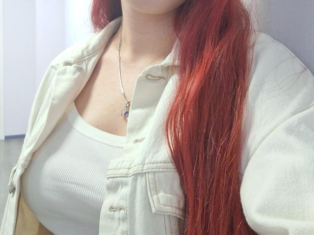 Triziya with red hair, white ribbed tank top, and unbuttoned beige jacket, showing her small boobs, wearing a necklace with a cross.