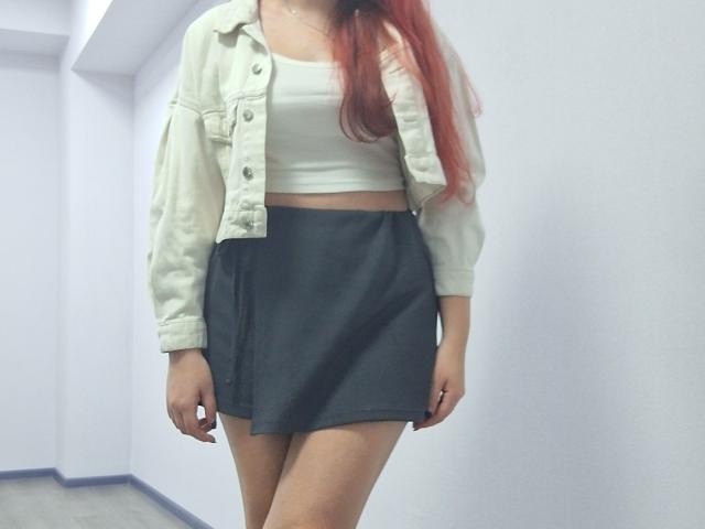 Triziya with red hair, wearing a white crop top, black mini-skirt, and light denim jacket, standing in a white-walled room. Her ass is partially visible.
