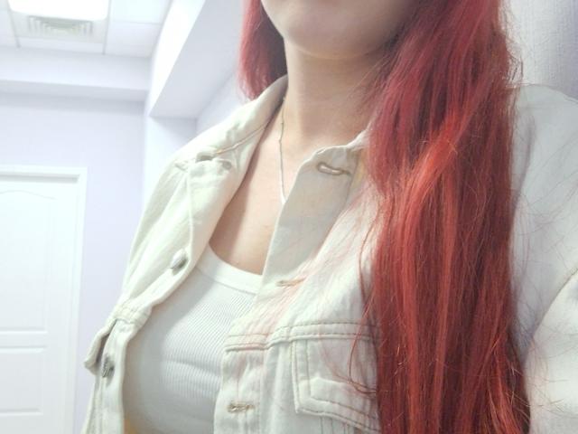 Triziya with long red hair, wearing a beige denim jacket over a white ribbed tank top, showing her boobs. She's standing in a bright room with a white door.