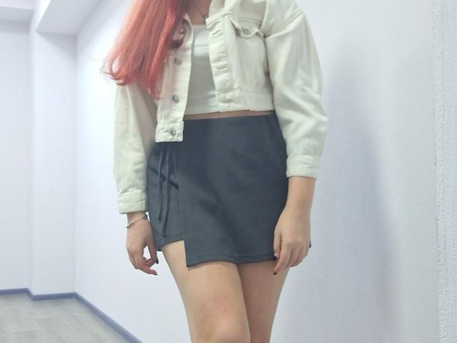 Triziya with red hair, white jacket, and black skirt, showing her ass and thighs, standing in a white-walled hallway.