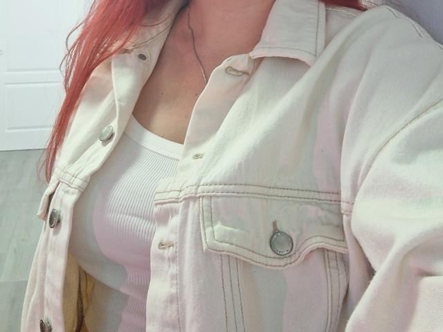 Triziya's pinkish-white denim jacket over a white ribbed tank top, showing her boobs. She has red hair, a silver necklace, and wooden floor in the background.