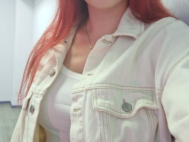 Triziya with red hair, wearing a white ribbed tank top and an open, light-colored denim jacket, showing her small tits. Her neck has a thin, silver chain.