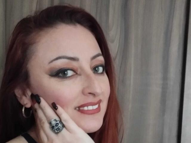 NoctisEye with red hair, black eyeliner, and red lipstick, smiling, touching cheek with black-nailed hand, wearing skull ring, hoop earrings, against gray curtain.