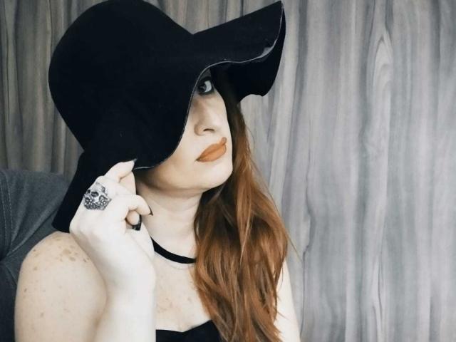 NoctisEye with long red hair, wearing a black hat covering one eye, has brown lipstick and a black ring, holding her hat's edge, showing freckled arms.