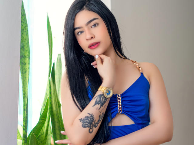 PaulinaFlame met lang zwart haar, een blauwe jurk, een gouden ketting, een tatoeage op haar rechterarm, rode lippenstift en een gouden horloge, die haar kin vasthoudt, naast een groene plant..