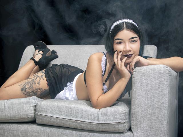 Eva Cullen, acostada en un sofá gris, vestida con lencería negra y encaje blanco, con tatuajes en el muslo, tacones altos negros y una diadema blanca, fumando con lápiz labial oscuro..