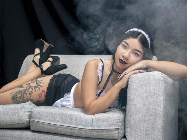 EvaCullen with black lipstick, lace bra, and choker, laying on gray couch, black high heels, tattoo on thigh, arm resting on arm, smoking, looking at camera.