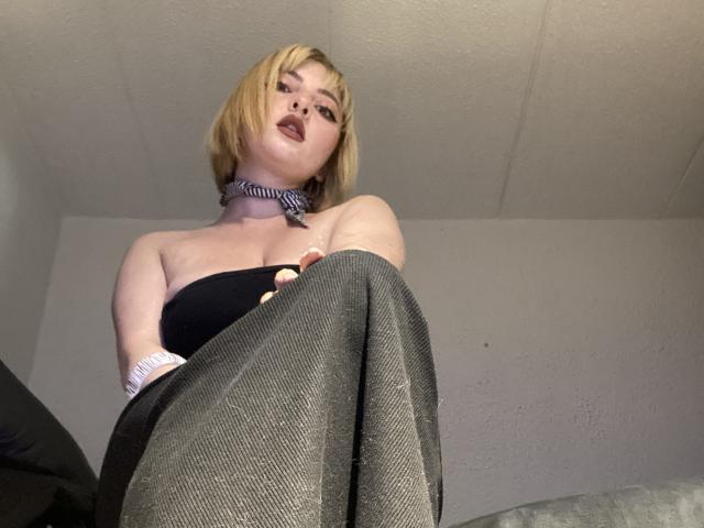 CocoVolkov con un corte de pelo rubio, un top negro sin tirantes, un collar, y leggings grises brillantes, con las piernas cruzadas, mirando hacia abajo, con la boca ligeramente abierta, lápiz labial oscuro, y el techo visible arriba..