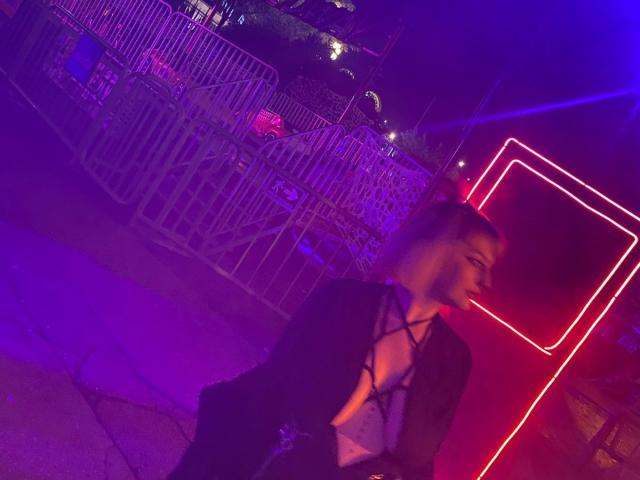CocoVolkov with dark strappy top, revealing cleavage, and black jacket, sitting in purple neon light, right side profile, short brown hair, background includes metal fence and neon sign.