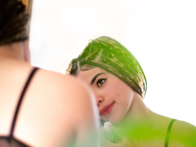 PasionSubmissive with green braids, looking at someone with black bra straps, slight smile, greenery in foreground, soft light, upper body, intimate moment, brown eyes, subtle makeup.