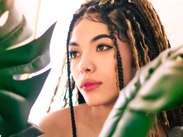 PasionSubmissive has curly brown braids, green eyes, pink lips, and a cute face. She's surrounded by green leaves in a bright, sunlit setting. Her ass is not visible.