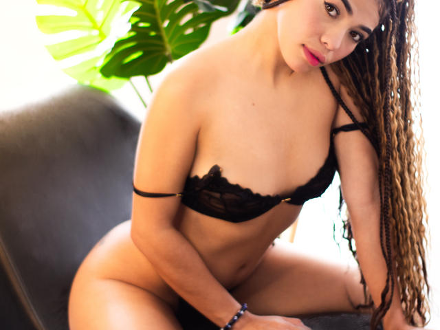 PasionSubmissive with long curly brown hair, wearing black lace bra, exposing her tits, sitting on a chair, looking at camera, pink lipstick, bracelet, green plant in background.
