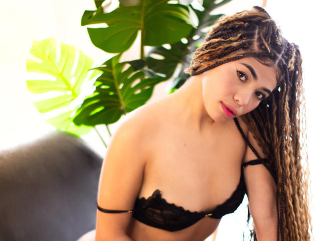 PasionSubmissive with long, curly brown hair wearing a black lace bra, leaning forward, showing small tits, and a cute, sexy look, with a green monstera plant in the background.