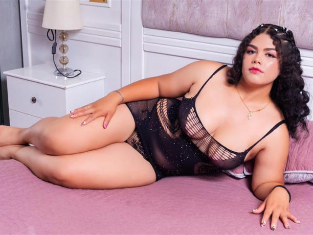 BrianaCleython with curly black hair, wearing a black sheer lingerie dress, lying on a pink bed, showing her large ass, small tits, and a confident expression.