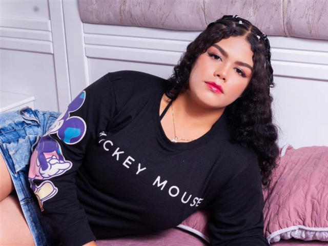 BrianaCleython with curly black hair, red lipstick, black "Cockey Mouse" sweatshirt, denim shorts, sitting on pink bed, looking at camera, slightly chubby, medium tits, confident expression.