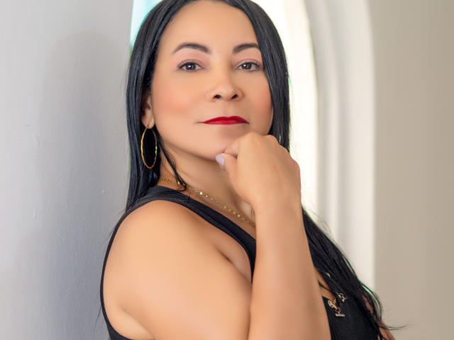 StefyDivine with black hair, red lipstick, and hoop earrings, touching her chin, wearing a black sleeveless top, showing her tits, looking at the camera with a slight smile.