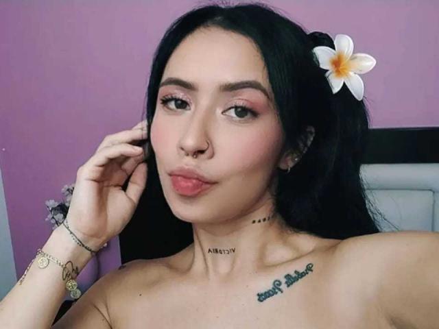 AlexaRosex with black hair, pink eyeshadow, nose ring, and flower in hair, pouting with hand near face, tattoos on neck and chest, bare shoulders, pink lipstick.