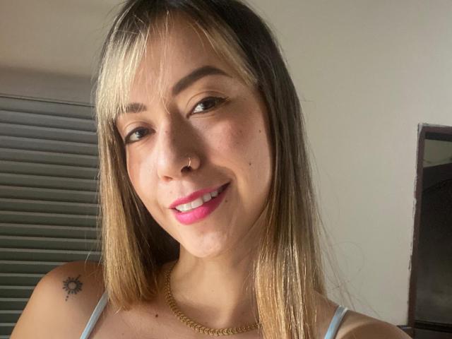 EmilyBonnett, avec des mèches blondes, souriante, rouge à lèvres rose, piercing au nez, tatouage de soleil sur l'épaule gauche, collier en or, portant un débardeur blanc, peau claire, poitrine moyenne, cheveux lisses..