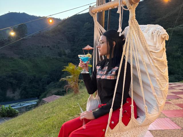 LizzeJoy with black hair, wearing a black top with colorful stripes and red pants, sips a blue cocktail on a rope swing, holding it in her right hand.
