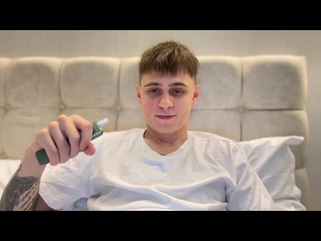 TylerGarner with short brown hair, wearing a white t-shirt, holding a green toothbrush, tattoo on right forearm, sitting up in bed, slightly smiling, soft lighting.