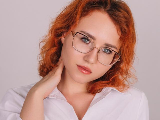 TeresaBirt with curly orange hair, wearing glasses, white shirt, hand on neck, blue eyes, pink lips, looking slightly to the side, soft expression.