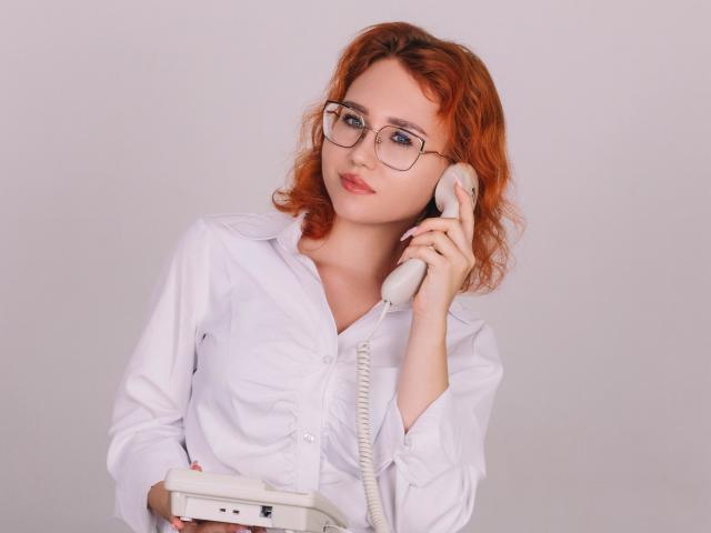 TeresaBirt with red curly hair, glasses, white button-down shirt, holding a landline phone to her ear, holding a white phone base with a cord in her other hand.