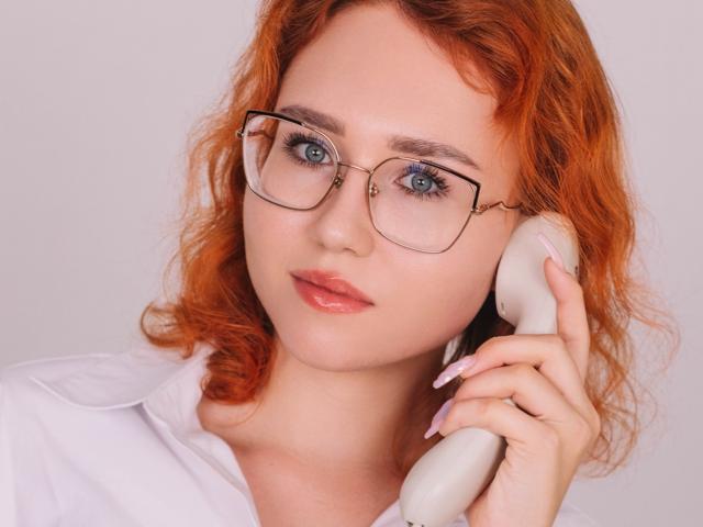 Teresa Birt, com cabelo ruivo encaracolado, óculos e batom rosa, segurando um telefone branco no ouvido, vestindo uma camisa branca, mostrando unhas longas e rosa..