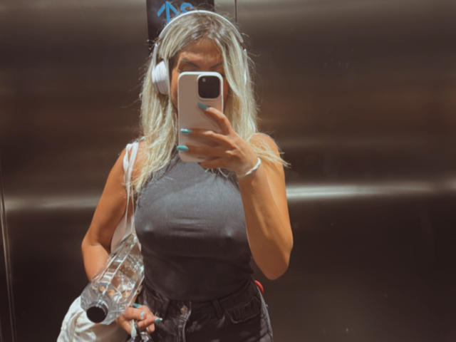 Lellie with blonde hair, gray sleeveless top, holding phone, wearing headphones, and blue nails, standing in elevator, holding clear plastic bag with her hand on her tits.