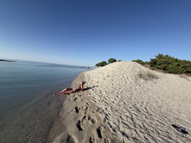 Lellie lies on a sandy shore with a small hill, her ass up, legs stretched, and arms above her head, under a clear blue sky, by calm water.