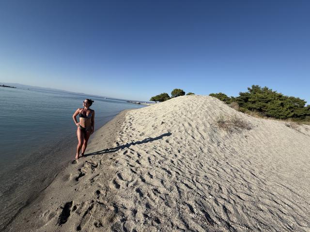 Lellie stands on a sandy hill with her brown bikini, hands on hips, showing her ass and small tits, near a clear blue water shoreline under a bright sky.
