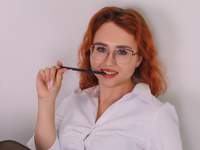 TeresaBirt with red curly hair, glasses, white shirt, holding blue pen in mouth, smiling, blue eyes, pink nails, soft light, slight blush, looking at camera.