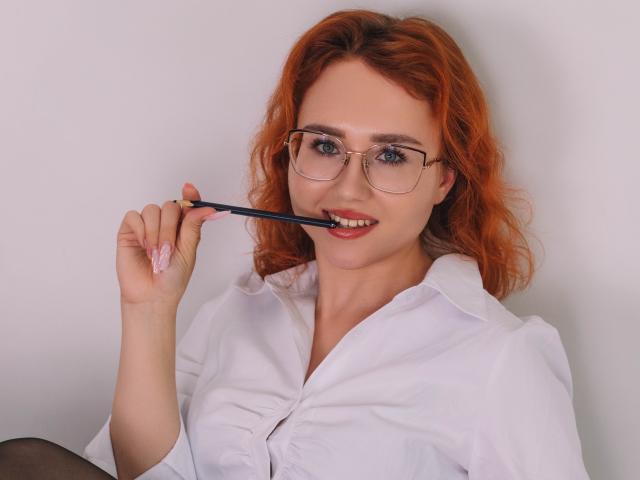 TeresaBirt with red hair, glasses, and white shirt, holding a black pen in her mouth, smiling. She has blue eyes and is wearing black tights.
