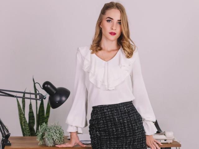 Eva Daly mit blonden Haaren, einer weißen, gerafften Bluse, einem schwarzen, strukturierten Rock, stehend neben einem Schreibtisch, einer schwarzen Lampe, einer grünen Schlange, einer kleinen Sukkulente, einer weißen Wand, rotem Lippenstift, ernstem Gesichtsausdruck..