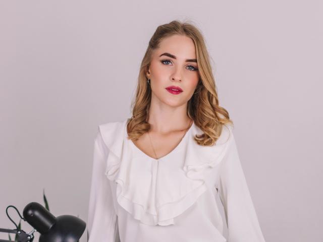 EvaDaly with blonde hair, blue eyes, red lipstick, wearing a white ruffled blouse, sitting in front of a black lamp on a white background.