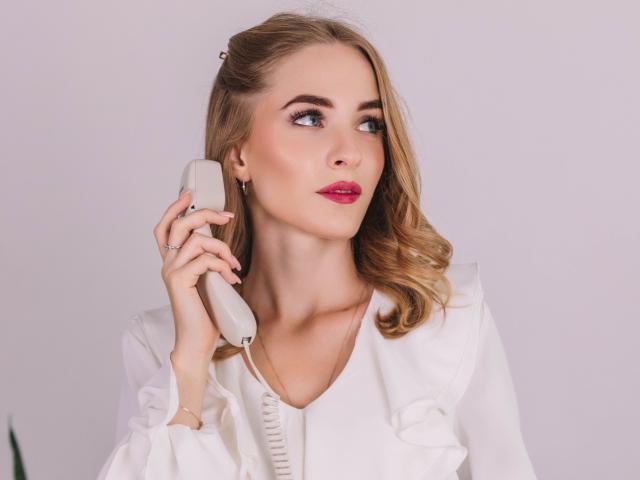 EvaDaly with blonde hair, holding a beige corded phone to her ear, wearing a white blouse, pink lipstick, and earrings, looking slightly to the side with a thoughtful expression.