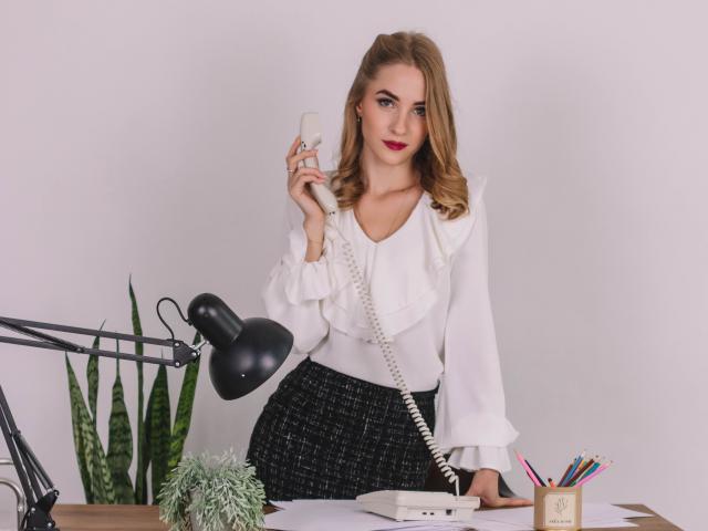 EvaDaly with wavy blonde hair, white blouse, black skirt, holding a phone, standing by a desk with a lamp, plant, and pencils, looking at the camera with red lipstick.