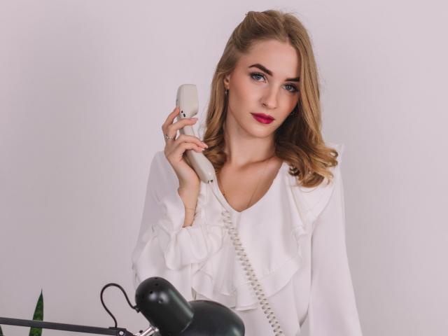 EvaDaly with blonde hair, white frilled blouse, holding a gray phone, sitting at a desk with a green lamp, looking confident with pink lipstick.