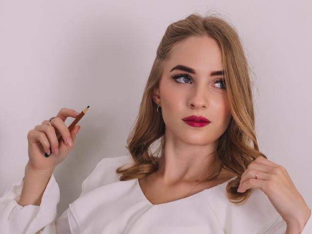 EvaDaly with blonde hair, blue eyes, red lipstick, white blouse, holding a brown eyeliner pencil, looking to the side, right hand touching hair, left hand holding eyeliner.