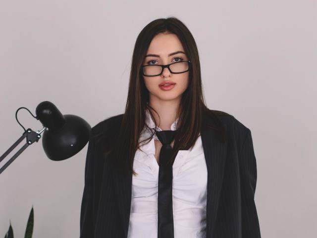ChristinaBliss, met een bril, draagt een zwart colbert met strepen, een wit shirt en een zwarte stropdas, heeft lang bruin haar en een lichte glimlach, en staat naast een zwarte lamp..